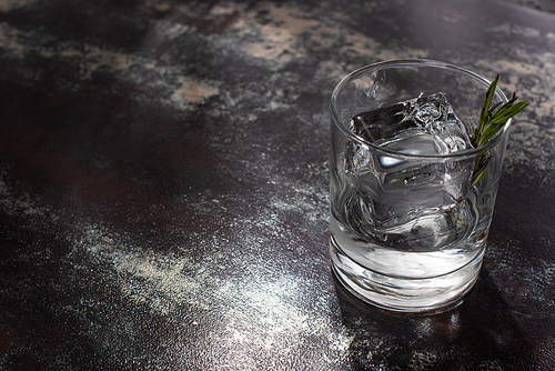 transparent glass with rosemary, ice cube and vodka on weathered surface
