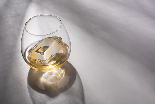 transparent glass with ice cube and whiskey on white table with shadow