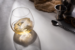 transparent glass with ice cube and whiskey on white table with shadow near cloth, spoon and jigger