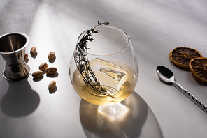 transparent glass with herb, ice cube and whiskey on white table with shadow near pistachios, spoon, dried citrus slices, jigger