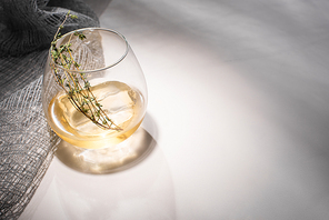 transparent glass with herb, ice cube and whiskey on white table with shadow near cloth