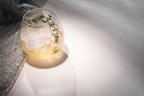 transparent glass with herb, ice and whiskey on white table with shadow near cloth