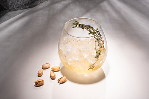 transparent glass with herb, ice cube and whiskey on white table with shadow near cloth and pistachios