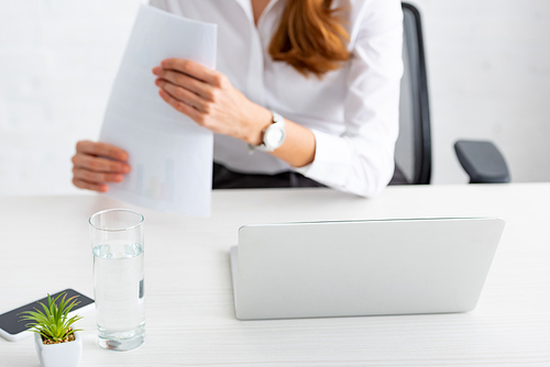 Selective focus of laptop and glass of water and businesswoman holding documents at table
