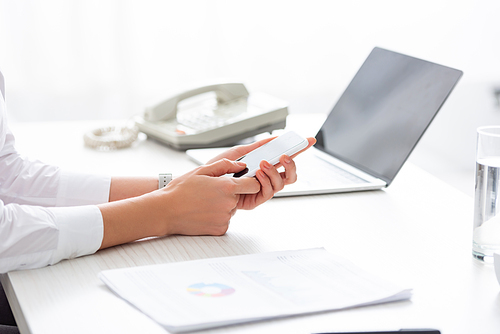 Cropped view of businesswoman using smartphone near documents and laptop on table