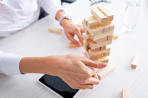 Cropped view of businesswoman playing blocks wood tower game near smartphone at table