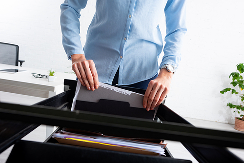 Cropped view of businesswoman taking folder with dossier from open cabinet driver in office