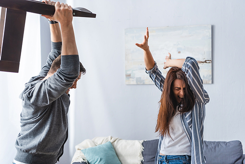 Abusive man holding chair near scared wife covering head at home