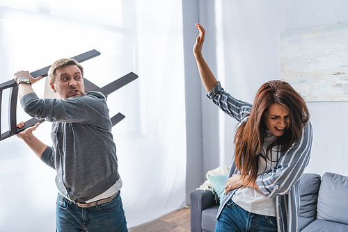 Angry man holding chair near frightened wife at home