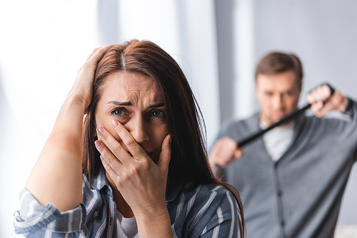 Worried woman with bruise on hand covering mouth near abusive husband with waist belt on blurred background