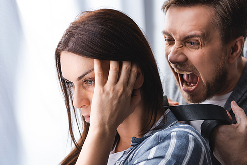Depressed woman standing near angry screaming husband with waist belt on blurred background