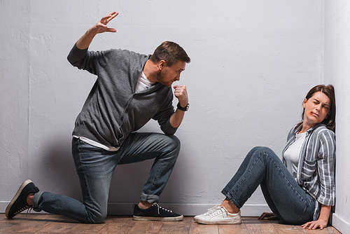 Angry man with hand in fist looking at wife with bruises on floor