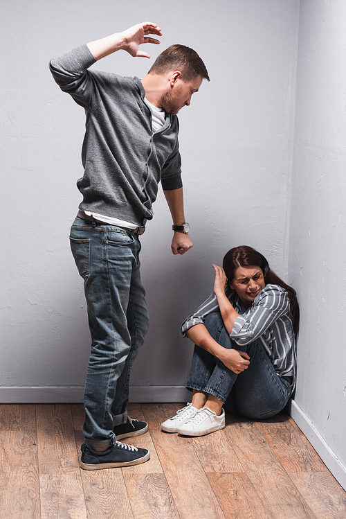 Abusive husband with hand in fist standing near scared woman with bruises on floor