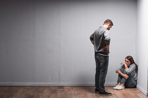 Abusive husband looking at depressed wife with bruises on face and hands on floor