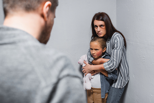 Woman with bruises hugging child with soft toy near walls and abusive husband on blurred foreground