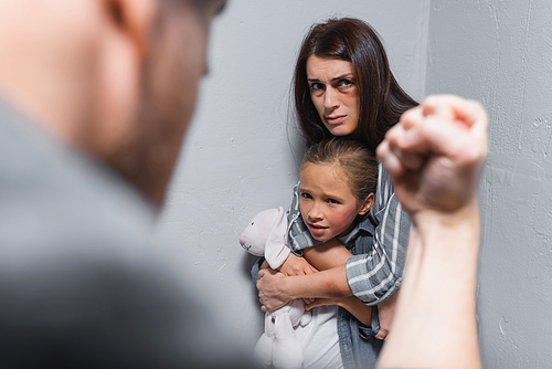Scared woman hugging daughter with bruise and soft toy near walls and husband showing fist on blurred foreground