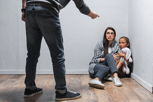 Child with soft toy and woman with bruises sitting on floor near abusive husband pointing with finger on blurred foreground