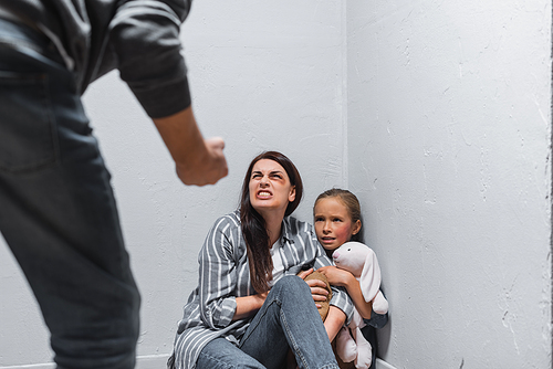 Angry woman with bruises on face sitting near child with soft toy and abusive man on blurred foreground