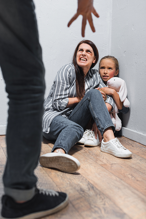 Screaming woman with bruises and kid with soft toy looking at abusive father on blurred foreground