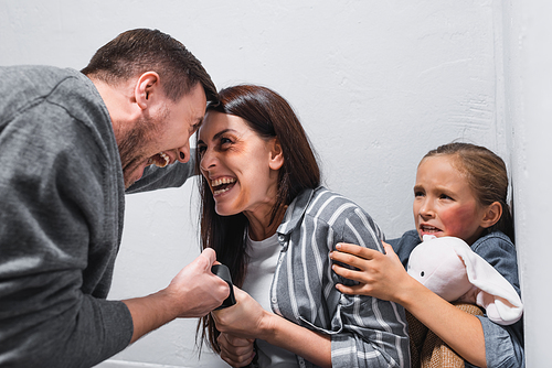 Screaming woman with bruises taking waist belt from husband hands near crying daughter