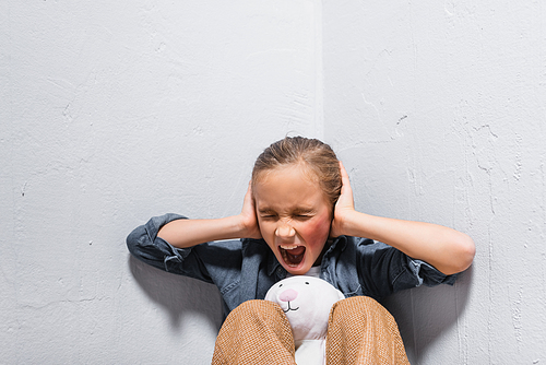 Screaming girl with hematoma on face covering ears near soft toy