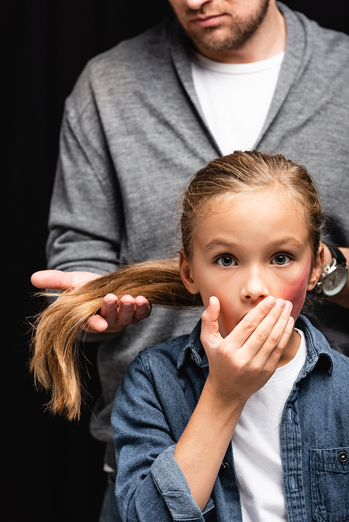 Scared kid with bruise on face standing near abusive father touching hair on blurred background isolated on black