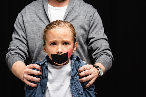Scared girl with bruise and adhesive tape on mouth standing near father on blurred background isolated on black