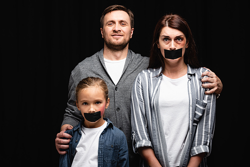 Smiling abuser hugging wife and daughter with bruises and adhesive tapes isolated on black