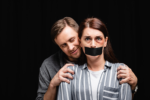 Smiling abuser hugging wife with hematoma and adhesive tape isolated on black