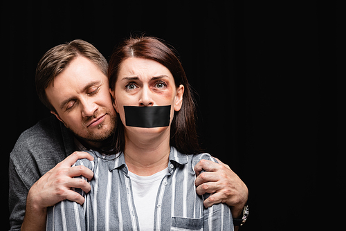 Scared woman with hematoma and adhesive tape on mouth standing near abuser isolated on black