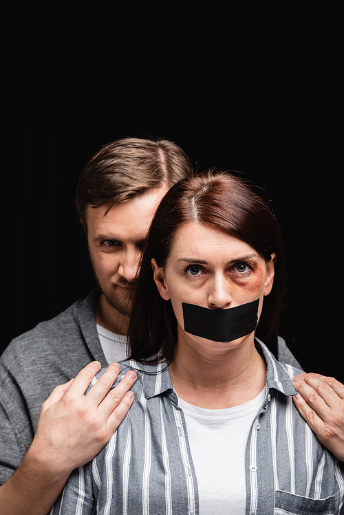 Abuser hiding behind wife with bruise and adhesive tape on mouth isolated on black