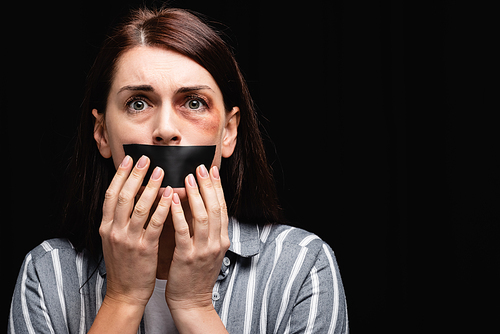 Scared woman in bruises touching adhesive tape on mouth isolated on black