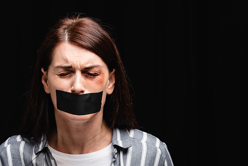 Crying woman with adhesive tape on mouth and bruise near eyes isolated on black