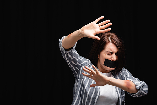 Scared woman with bruises and adhesive tape showing stop gesture isolated on black