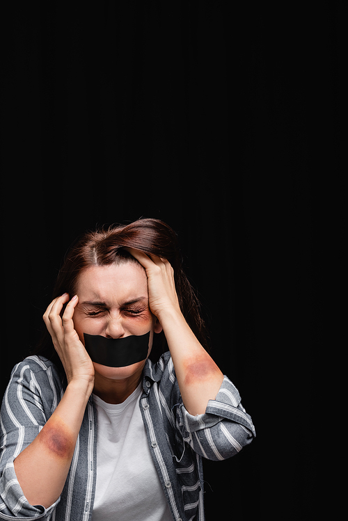 Depressed woman with bruises and adhesive tape on mouth isolated on black