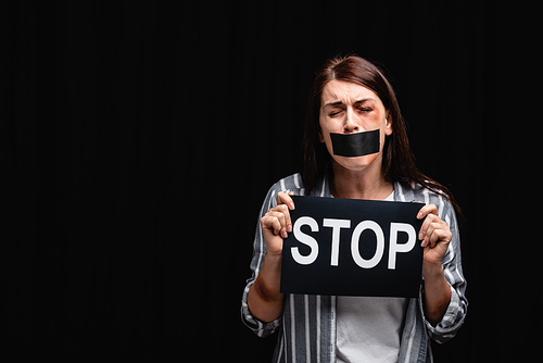 Offended woman with adhesive tape on mouth and bruises holding card with stop lettering isolated on black