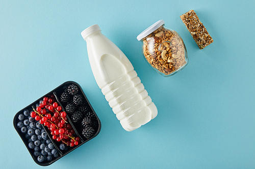 Top view of container with berries, bottle of milk, jar of granola and cereal bar on blue background