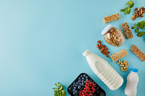 Top view of container with berries, bottles of yogurt and milk, jar of granola, nuts, cereal bars on blue background