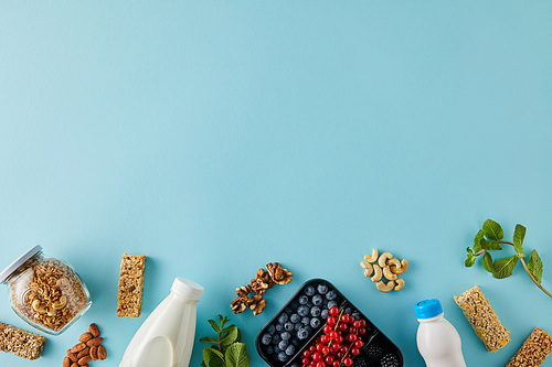 Top view of container with berries, bottles of yogurt and milk, jar of granola, nuts, cereal bars and mint on blue background