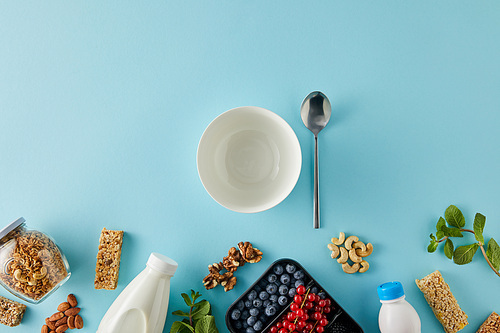 Top view of container with berries, bottles of yogurt and milk, jar of granola, nuts, cereal bars, bowl, spoon on blue background