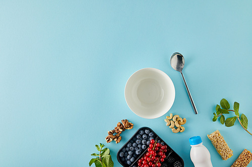 Top view of empty bowl with spoon, container with berries, bottle of yogurt, nuts, cereal bars, mint on blue background