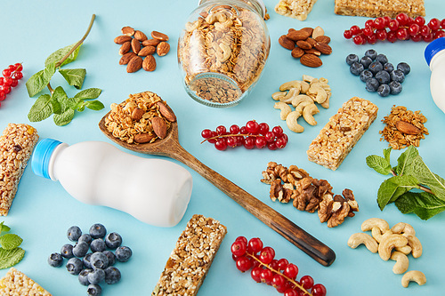 Jar of granola, bottles of yogurt, wooden spatula and berries with nuts around on blue background