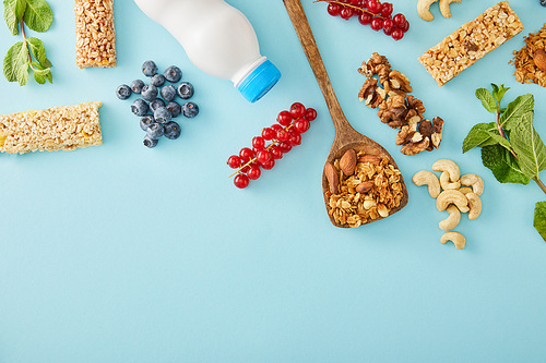 Top view of bottle of yogurt, berries, mint, nuts and cereal bars on blue background