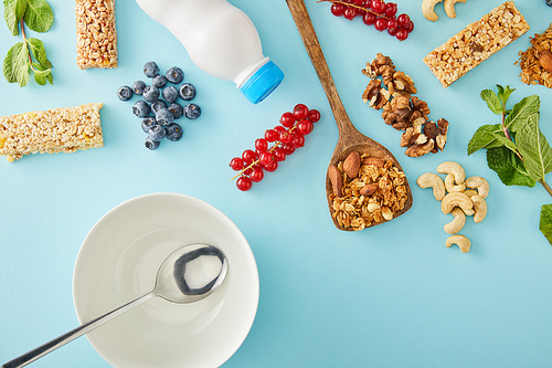 Top view of spoon inside empty bowl with berries, mint, nuts, bottle of yogurt and cereal bars on blue background