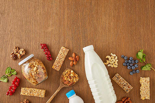 Top view of bottles of milk and yogurt with berries, mint, nuts, cereal bars and jar of granola on wooden background