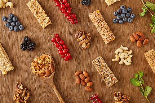 Top view of food composition of berries, nuts, cereal bars, mint and spatula with granola on wooden background