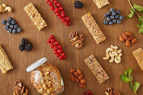Top view of food composition of berries, nuts, cereal bars, mint and jar of granola on wooden background