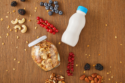 Top view of berries, nuts, jar of granola and bottle of yogurt on wooden background