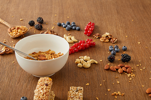 Bowl with granola next to berries, nuts and cereal bars on wooden background