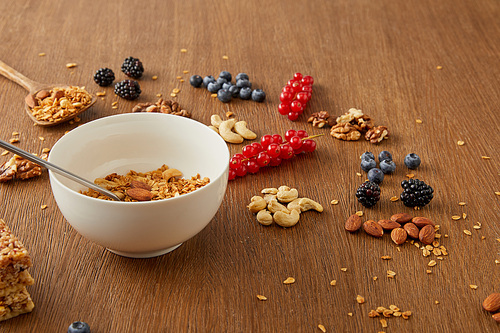 Bowl with granola next to blueberries, redcurrants, walnuts, almonds, cashews on wooden background